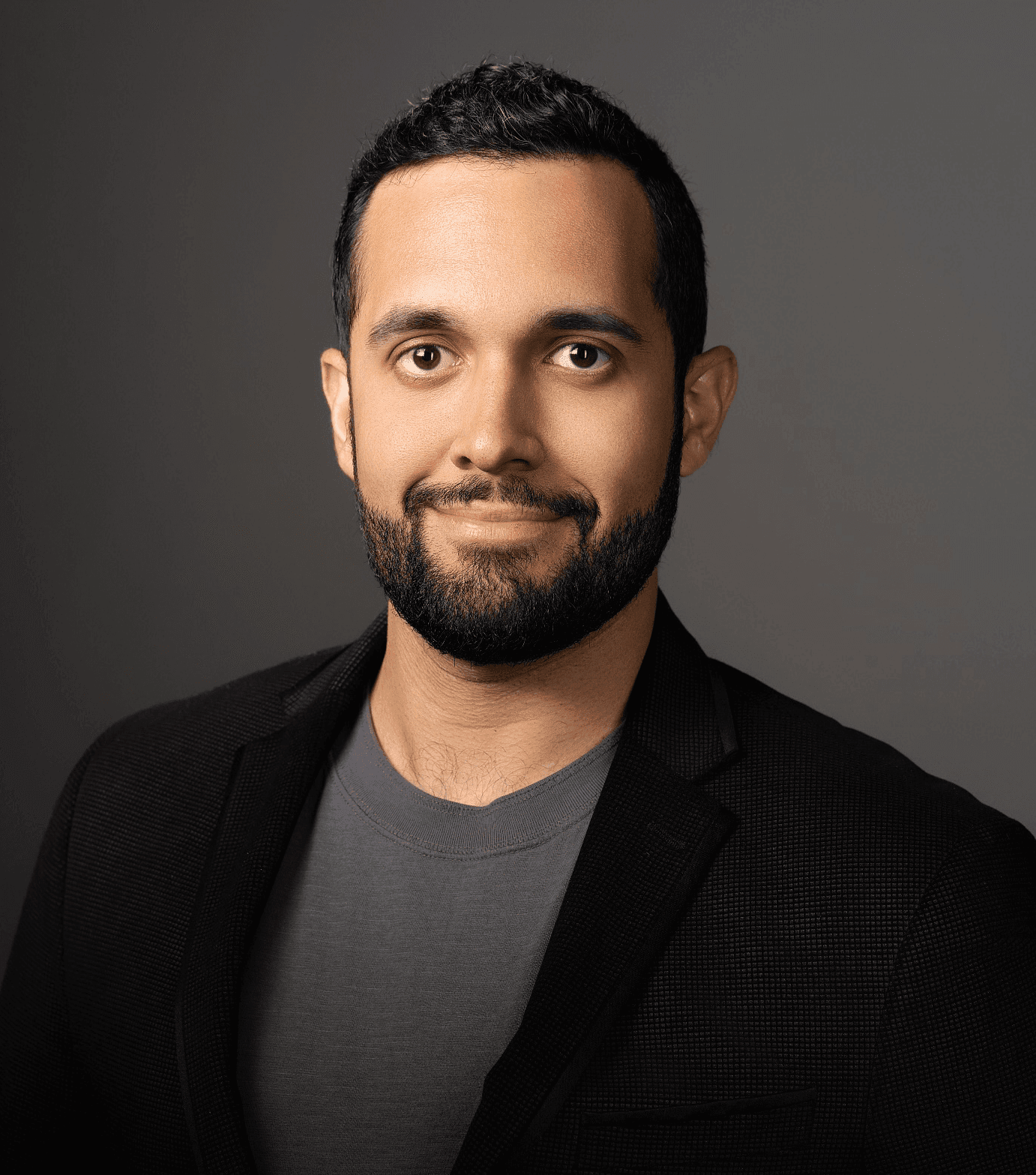 Person wearing a dark blazer and a gray shirt against a dark background.