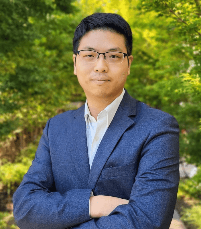 Person in a blue suit with arms crossed, standing outdoors with green trees in the background.